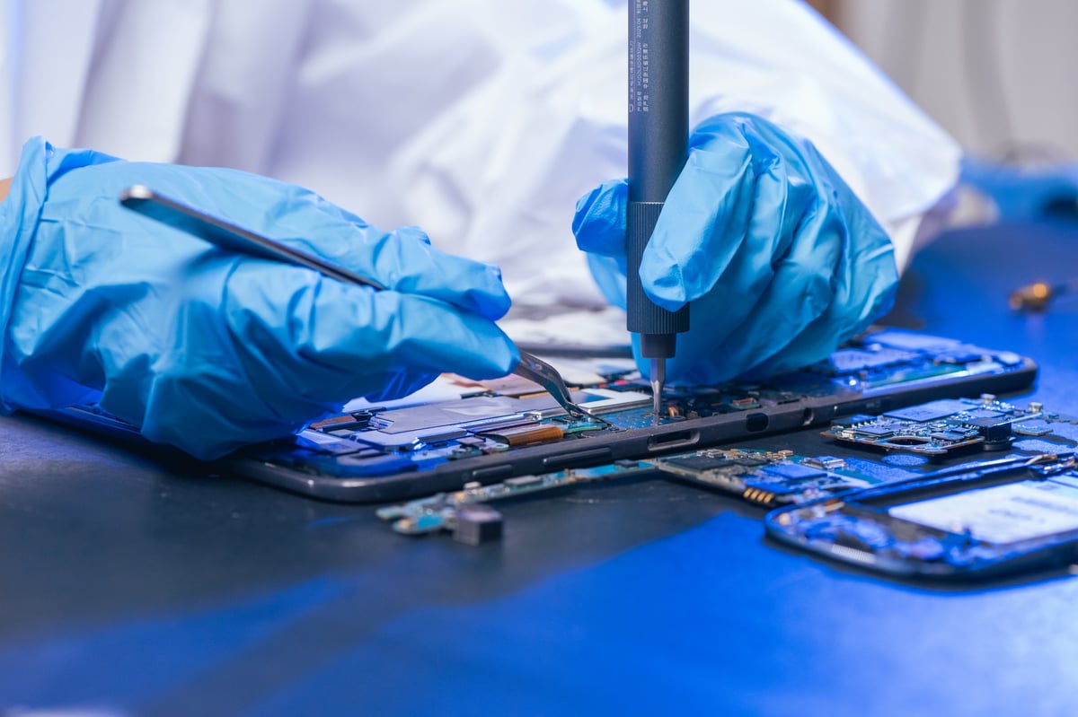 young female electronics technician carefully repairs a smartphone at her workstation. She uses professional precision screwdrivers and tweezers under a bright workspace light.